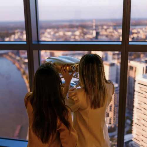 1vise_od_panorame_-1-scaled-2 Two girls observe the panoramic view of Belgrade and the river through a telescope from the Belgrade 360 Deck at dusk.