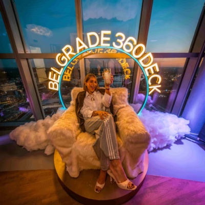 Gošća pozira u photo corner-u Belgrade 360 Deck-a sa neonskim natpisom i noćnom panoramom Beograda u pozadini.