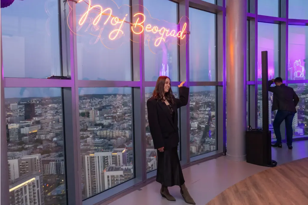 Devojka pozira na Belgrade 360 Deck-u ispred svetlećeg natpisa „Moj Beograd“, sa panoramskim pogledom na grad.