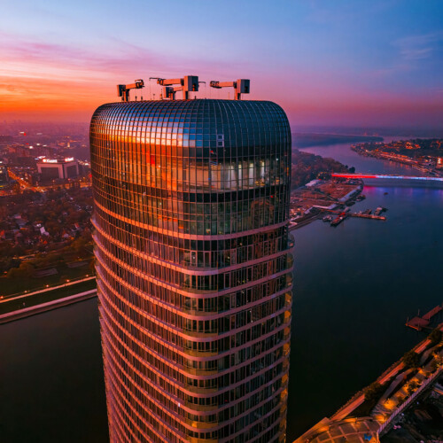 Pogled iz vazduha na vrh Kule Beograd u zalasku sunca. Narandžasto nebo i svetla grada reflektuju se na staklenoj fasadi.